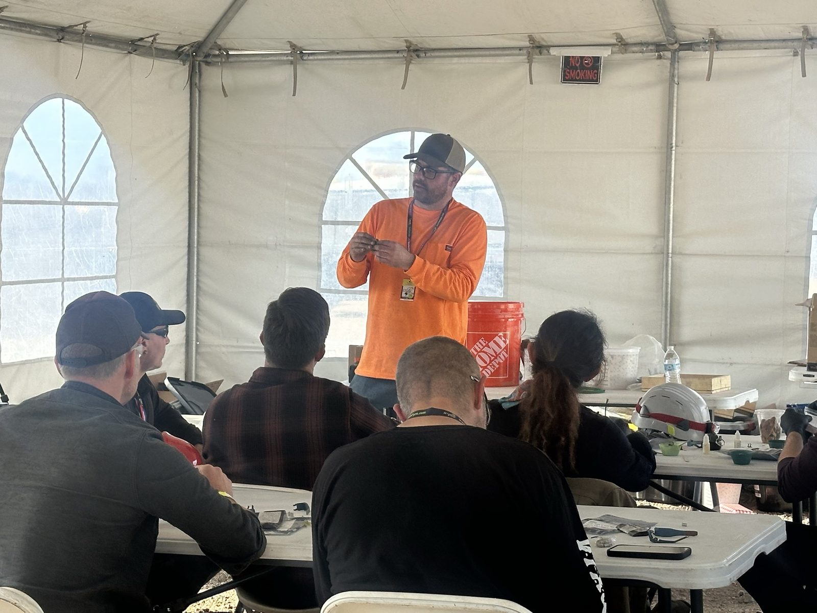 An instructor in an orange shirt and cap stands at the front of a tent classroom holding a small component, speaking to students seated at tables.